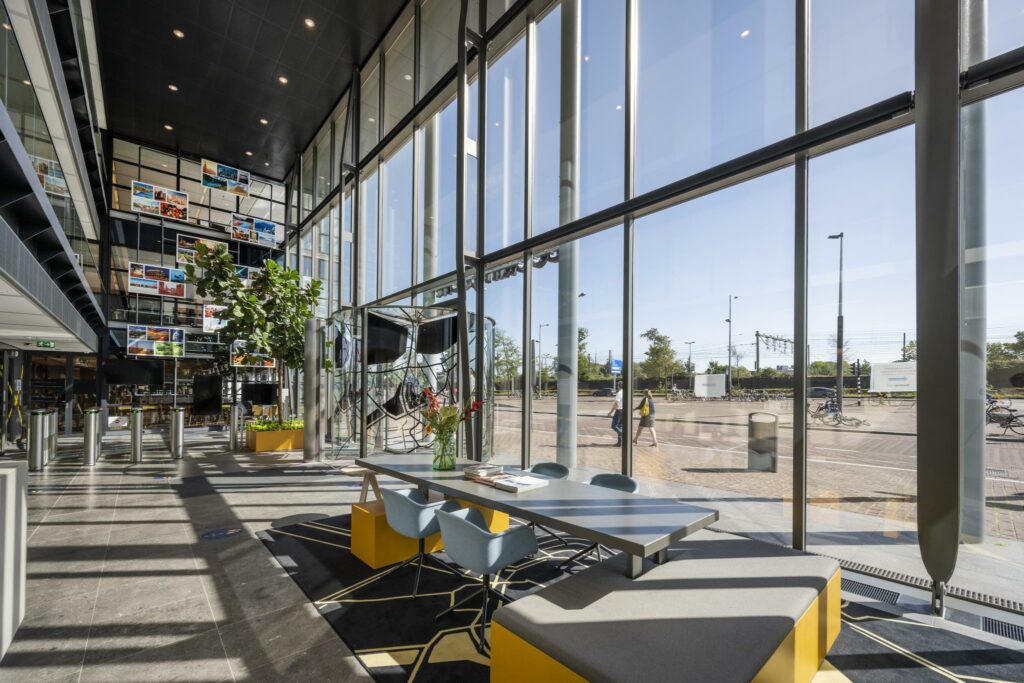 Moderne, lichtovergoten lobby aan de Piet Heinkade met grote ramen, kleurrijke zitplekken en uitzicht op het spoor.